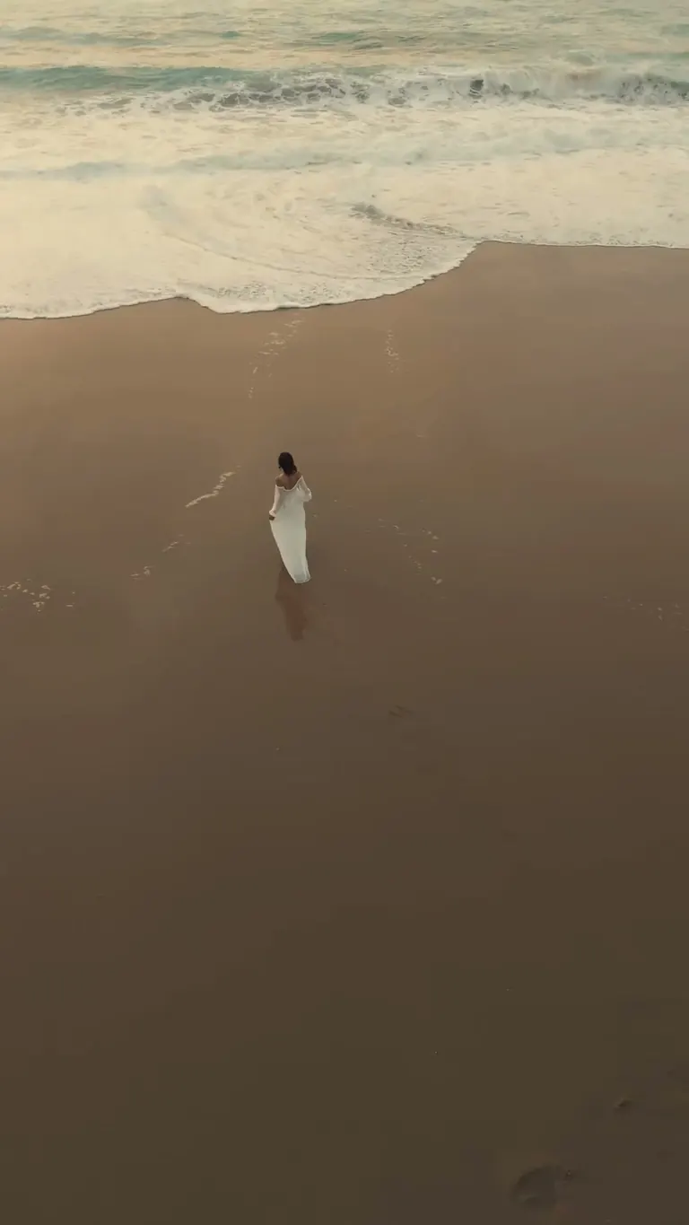 Seen from above, a bride walks along the wet sand at the water’s edge