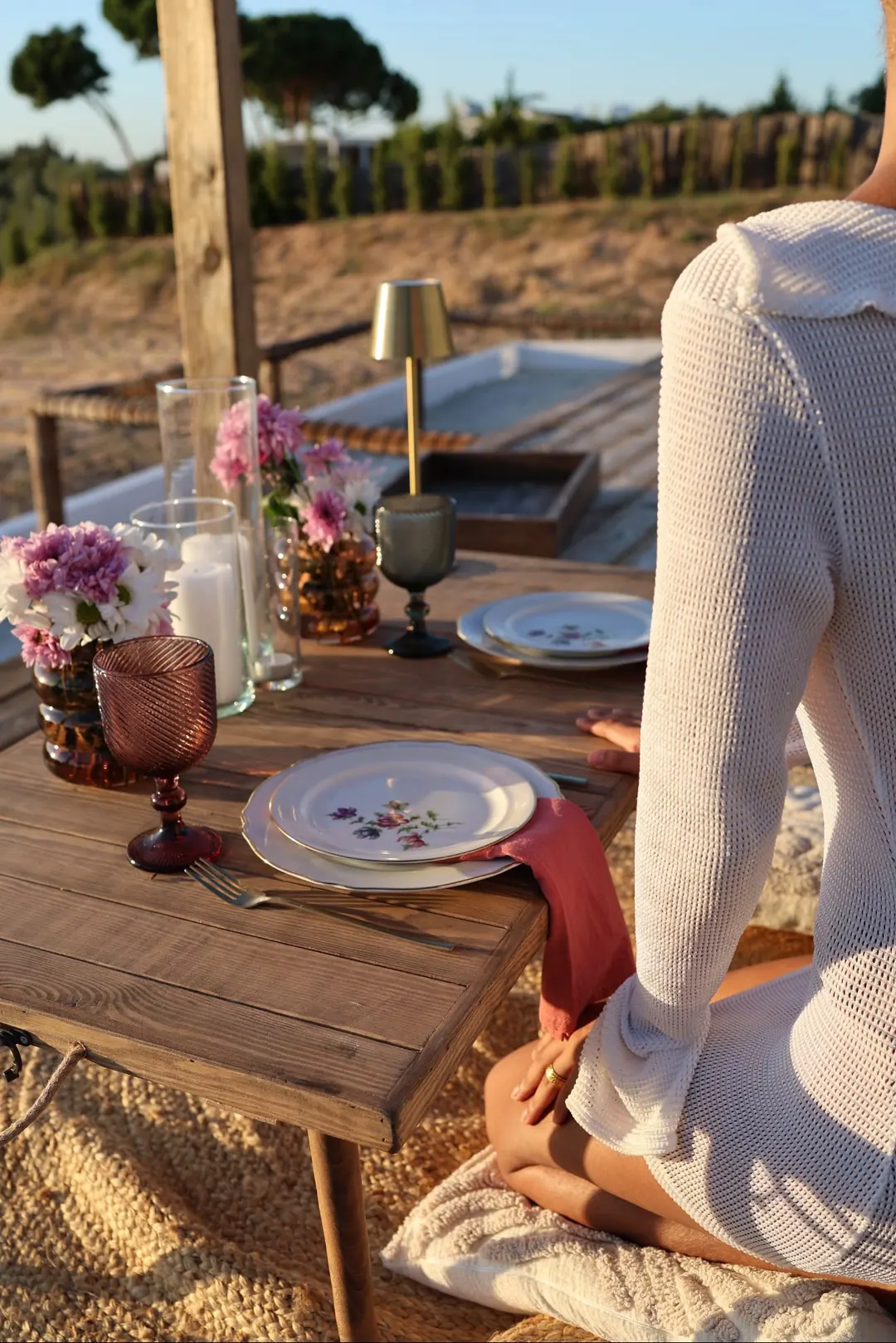 A wooden table by the pool with pink flowers, coloured glassware and painted plates
